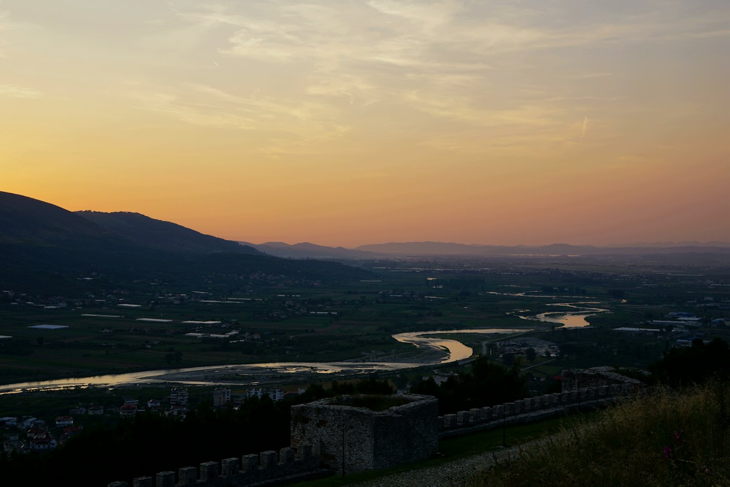Valle al tramonto con fiume che riflette le sfumature del cielo e montagne in lontananza.
